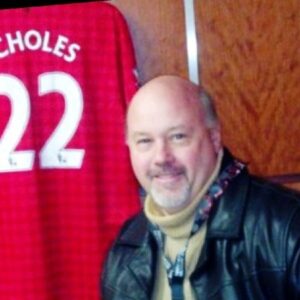 A Caucasian middle-aged man is smiling in a photo with a red jersey in the background.