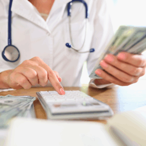 Doctor counting money while sitting at medical clinic desk with calculator.