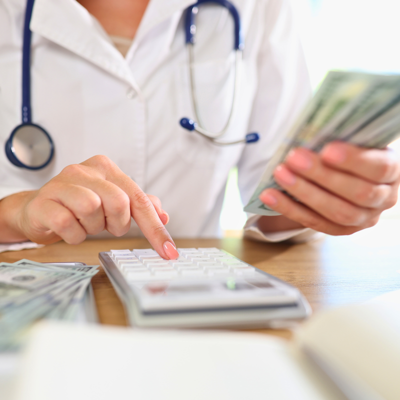 Doctor counting money while sitting at medical clinic desk with calculator.