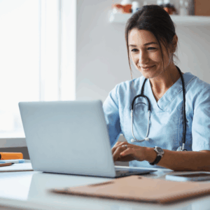 A woman wearing blue scrubs is working on a laptop, appearing engaged in her task.
